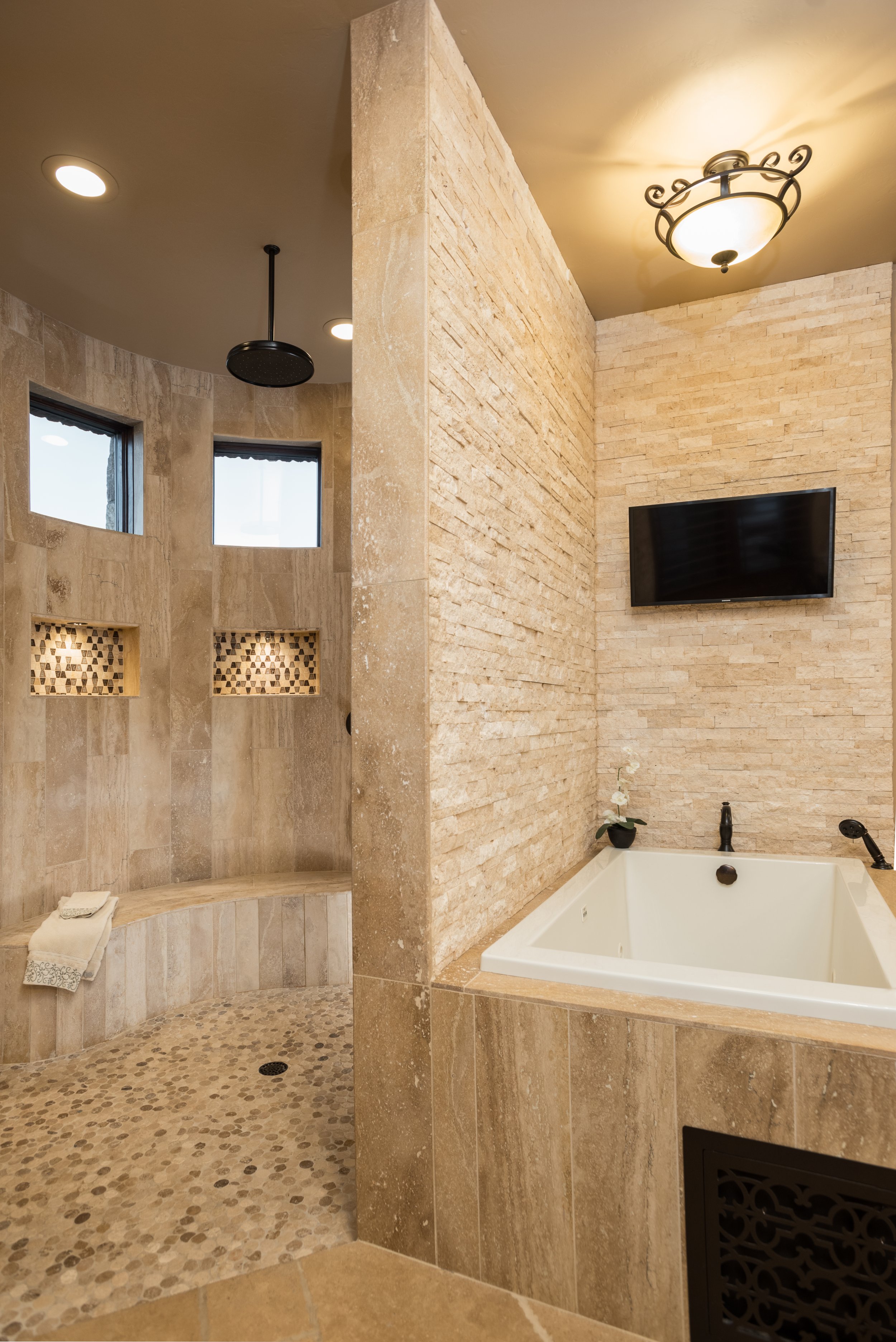 Luxury bathroom in Tucson with stone tiles, large shower area with built-in shelves, textured wall, recessed lighting, bathtub, and mounted TV.