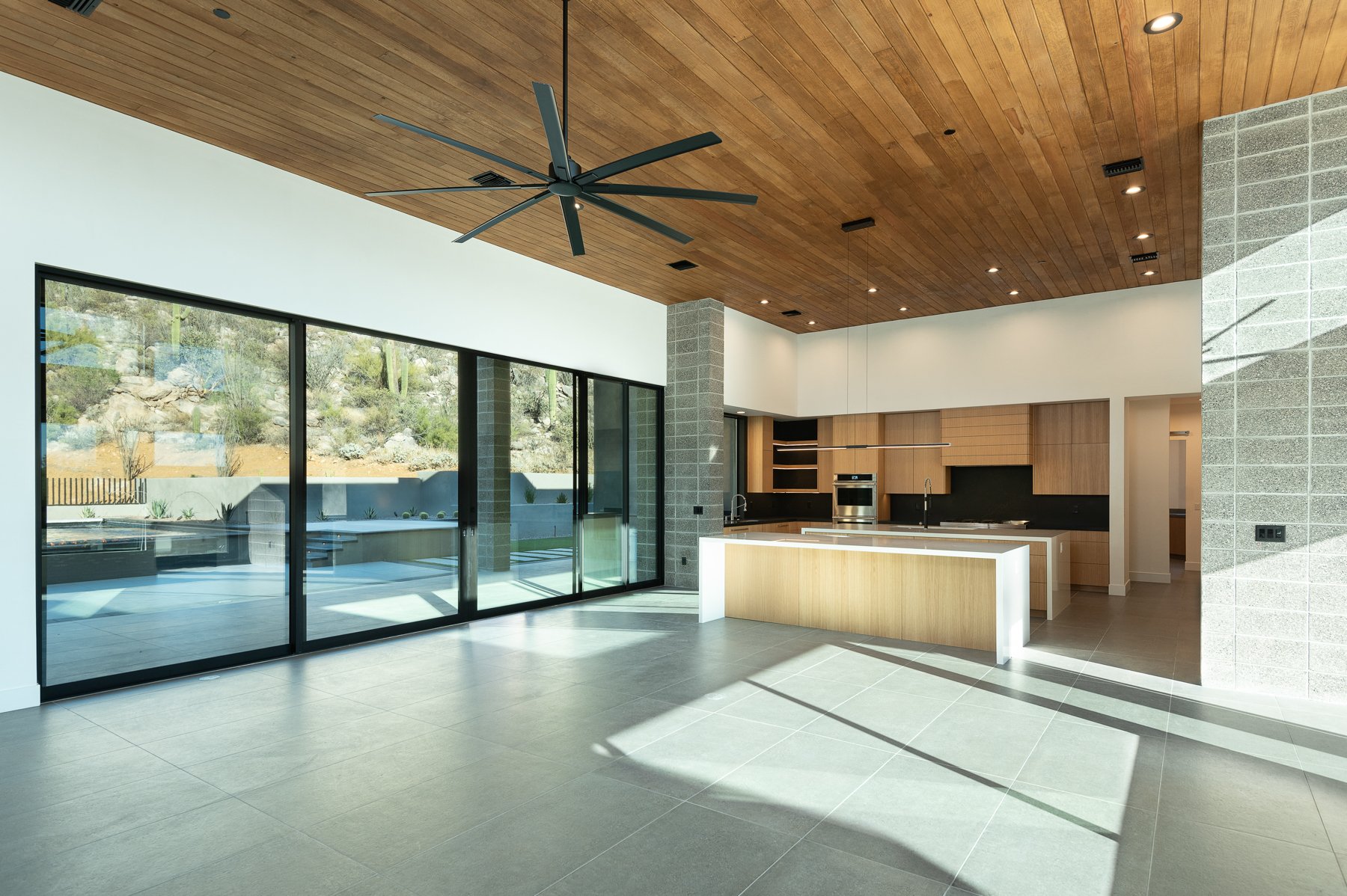 Modern open-plan kitchen and living area with large windows, wood-paneled ceiling, and ceiling fan. Outdoor view of desert landscape.