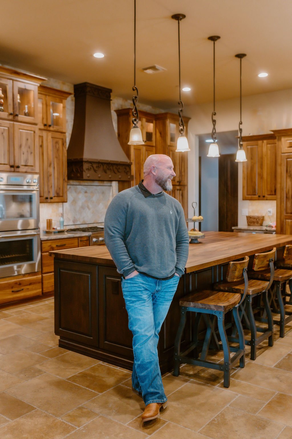 A man standing in a modern kitchen with wooden cabinets, a large center island, pendant lighting, and stainless steel appliances.