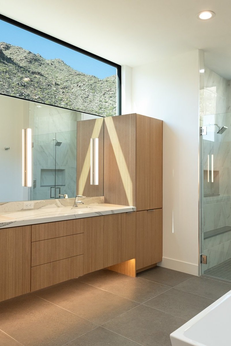 Modern bathroom with wooden cabinets, large mirror, marble countertop, glass shower, and mountain view through large window.